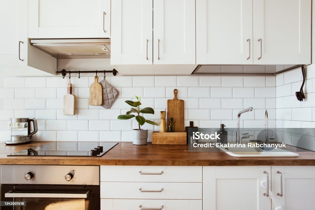 Hanging kitchen featuring brass utensils and chef accessories, complemented by a white tiled wall and wooden tabletop, with a green plant in the background.