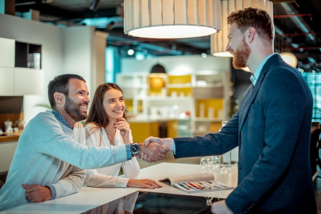 Three people smile as they shake hands at a bright showroom counter, with a color swatch book open on the table in front of them.